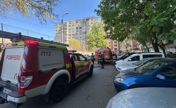 Firefighters in turnout gear work near red fire trucks in a residential parking lot beside apartment buildings.