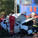 White car severely damaged after collision with a blue train, emergency responders in red and yellow gear assisting on the tracks.
