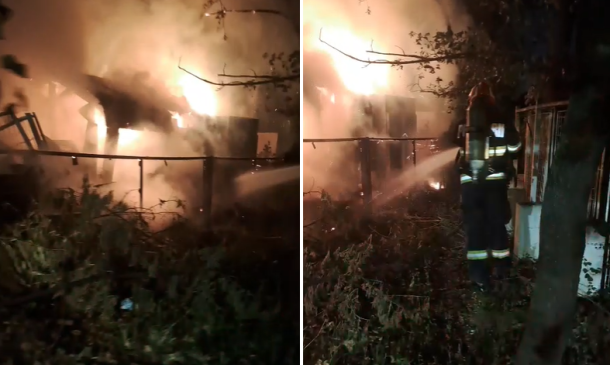 Nighttime yard fire with flames and smoke; a metal fence and debris, firefighters spraying water toward the blaze.