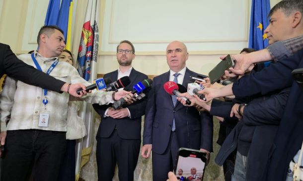 Bald man in a suit addressing reporters with microphones at a press conference, journalists surrounding him.