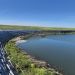 Curved wall of stacked concrete bags along a calm water body under a clear blue sky and grassy embankment beyond.