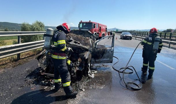 Firefighters in turnout gear extinguish a burned car on a highway, smoke rising, with a red fire engine in the background and a wet, reflective road surface.