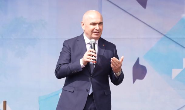 Man in a dark suit speaks into a handheld microphone on a stage with a blue abstract backdrop.