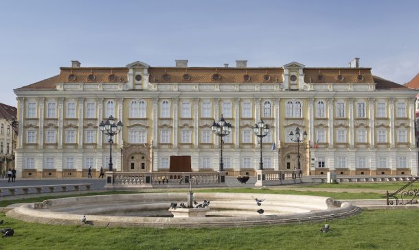 Facade of a grand neoclassical building with arched windows and a central entrance, in front of a circular fountain and pigeons in a park setting
