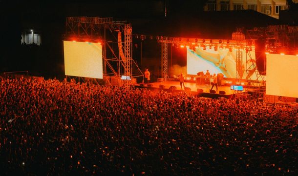 Outdoor night concert with a dense crowd facing a brightly lit stage and two large screens.