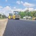 Two yellow road rollers compacting fresh asphalt on a rural street, with parked cars and workers in the background.