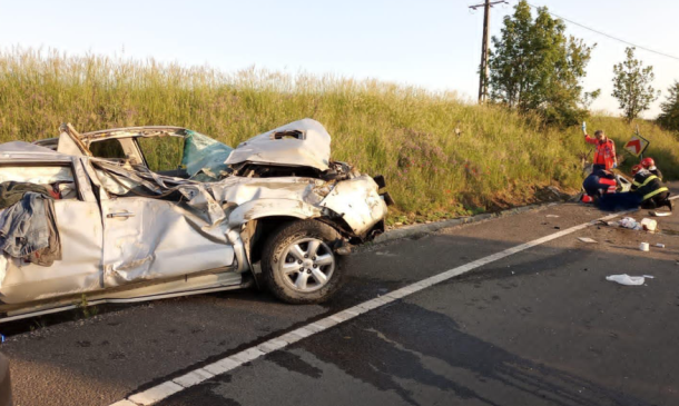 Wrecked white sedan after a severe crash on a road with emergency responders tending to a person nearby on the grass verge to the right of the scene.