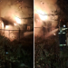 Nighttime yard fire with flames and smoke; a metal fence and debris, firefighters spraying water toward the blaze.